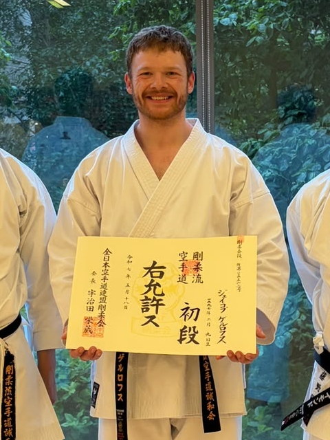 Jake smiling in his karate gi, holding his shodan (first-degree black belt) certificate.