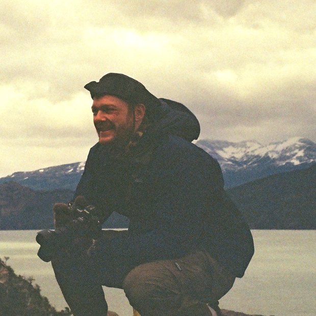Jake crouching with a camera on a lakeshore in Patagonia, snow-capped mountains behind.